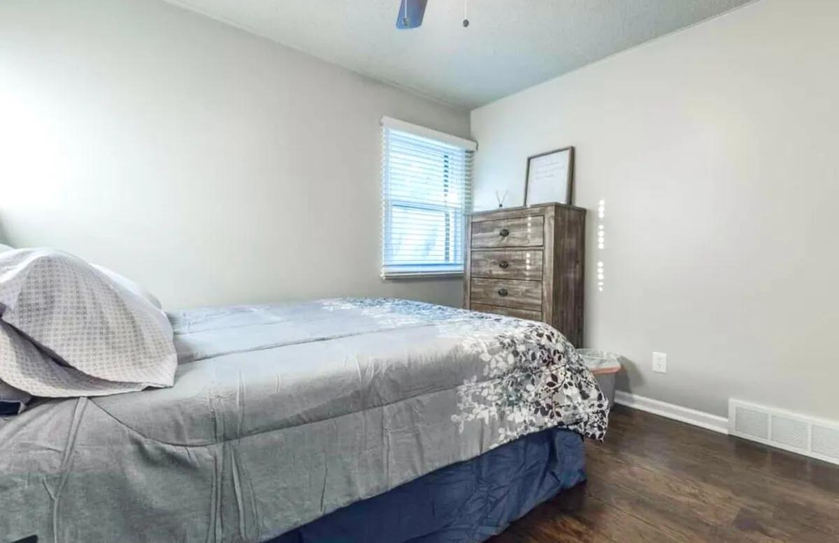 This image features a serene bedroom with a comfortable bed, a dresser for storage, and a window allowing in gentle natural light. The bedding and decor evoke a tranquil atmosphere, ideal for relaxation and restful sleep.