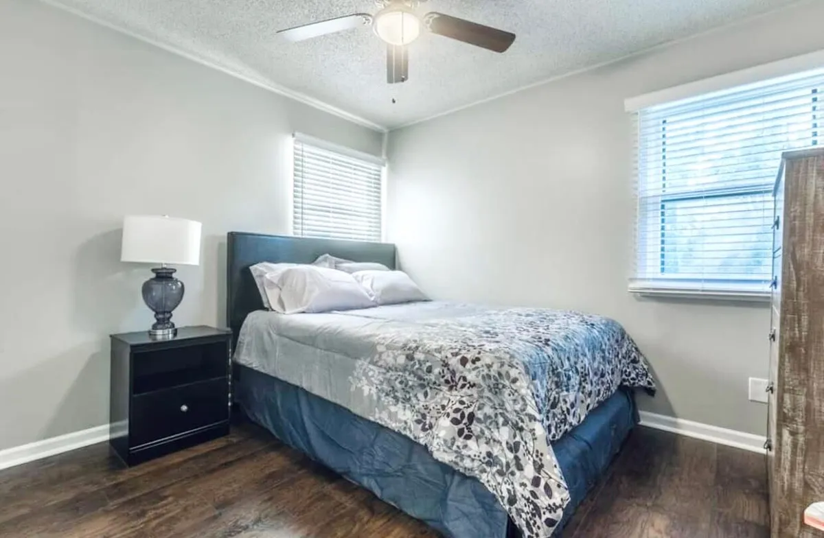 A tranquil bedroom showcasing a neatly made bed, a nightstand with a lamp, and large windows that fill the room with natural light. The space is decluttered and restful, perfect for napping or winding down after a busy day.