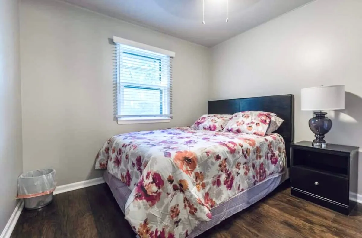 A cozy bedroom scene with floral bedding, a nightstand with a reading lamp, and a window dressed with blinds. The room exudes warmth and invites relaxation, perfect for quiet evenings or restful nights.