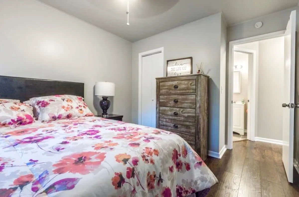 A cozy bedroom featuring a bed with inviting floral bedding, a classic wooden dresser, and a soft lamp that casts a warm glow. Gentle natural light streams in through a nearby window, creating a peaceful, restful atmosphere—perfect for unwinding after a productive day.