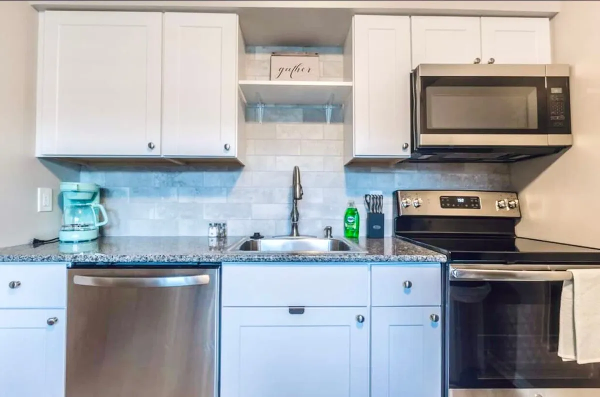 A bright, open kitchen featuring stainless steel appliances, ample granite countertop space, white cabinetry, and a kitchen sink. A decorative sign adds a touch of warmth, hinting at the kitchen's role as a hub for family gatherings and meal prep.