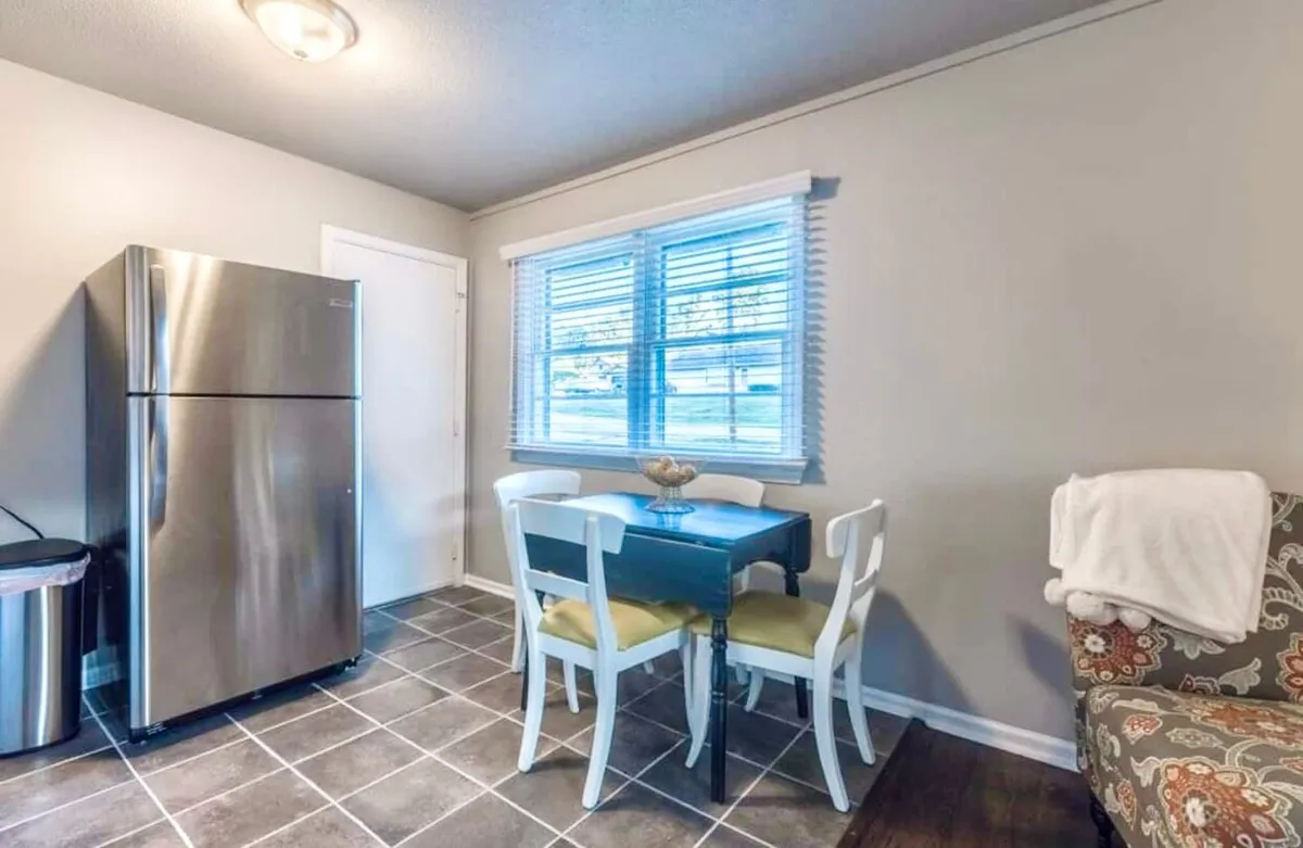 An inviting kitchen scene with a stainless steel refrigerator, small dining table for casual meals, a window with blinds that lets in soft light, and a textured armchair. The setting feels welcoming and functional for both family gatherings and quiet moments.