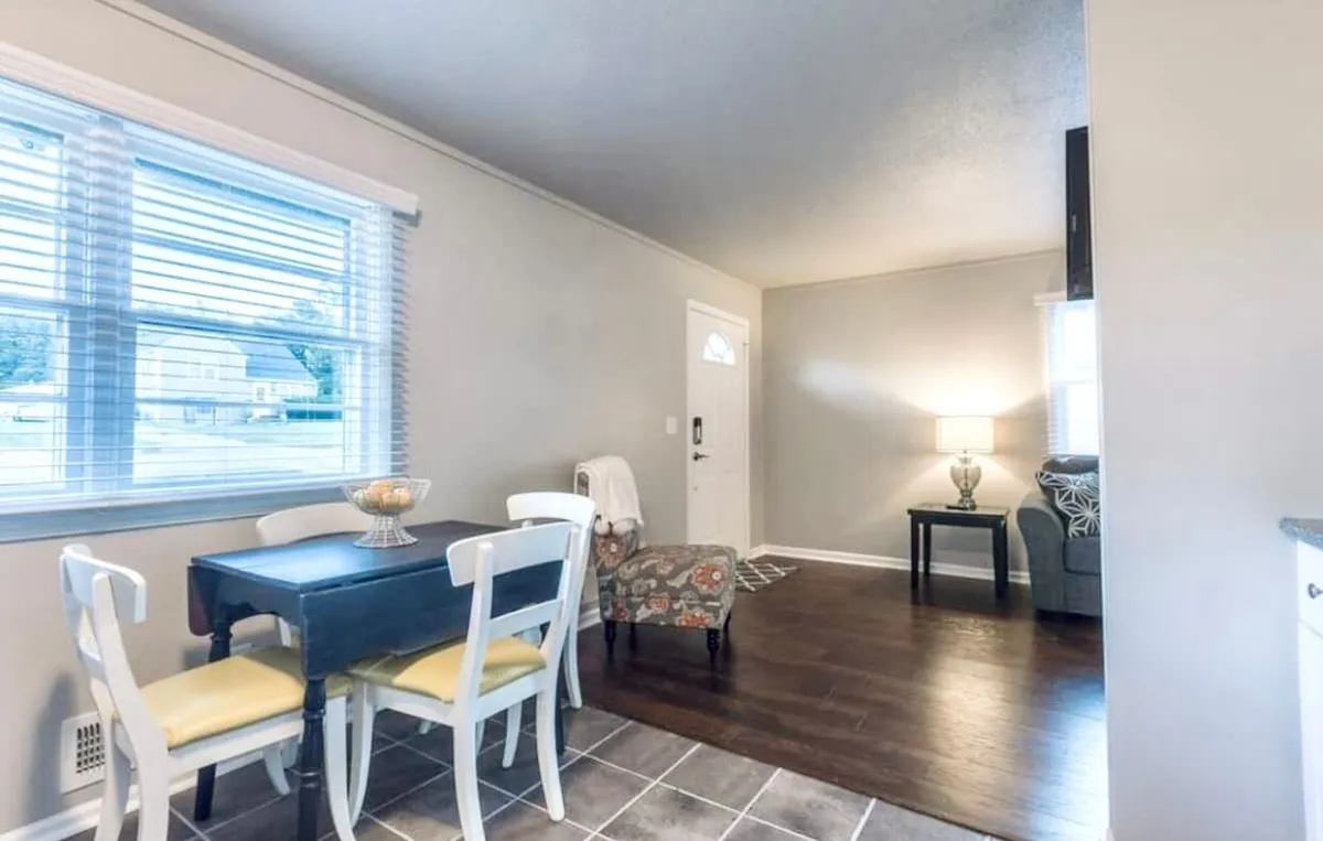 A cozy dining area featuring a large dining table, armchair, doorway, and a warm lamp. Sunlight fills the space, making it a cheerful spot for shared meals or conversations, with enough seating for family and friends.
